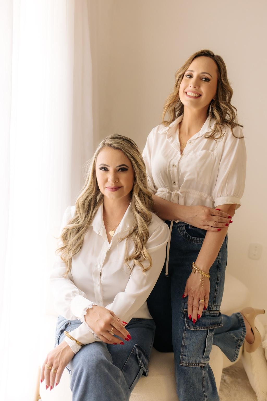 As duas proprietárias da SU Aromas e Artes, sorrindo, vestindo camisas brancas e calças jeans, em um ambiente claro